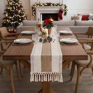 Peut inclure: Une table à manger en bois dressée pour un repas de fête. La table est ornée d'un chemin de table blanc et beige avec des franges, des assiettes, des verres, des bougies, des pommes de pin et un poinsettia. Un sapin de Noël et une cheminée sont visibles en arrière-plan.