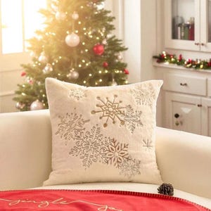 Pu&ograve; includere: Cuscino decorativo color crema con ricami di fiocchi di neve di varie dimensioni e filo dorato metallico. Il cuscino &egrave; su un divano bianco, con un albero di Natale sullo sfondo.