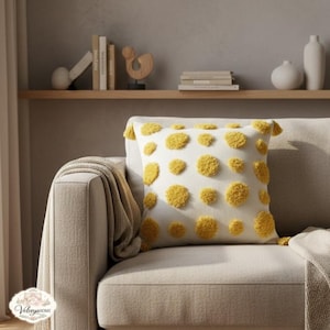 Peut inclure: Un coussin carré avec un fond blanc et de grands pois jaunes touffetés. Le coussin a des glands jaunes à chaque coin et est placé sur un canapé beige. Le coussin fait partie d'une collection de décoration intérieure.