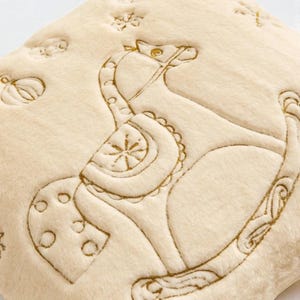 Pu&ograve; includere: Cuscino in peluche color crema con un design di cavallo a dondolo ricamato con filo dorato. Il cuscino presenta ulteriori dettagli ricamati di una zucca, stelle e altre forme. Il materiale in peluche offre un aspetto morbido e strutturato.