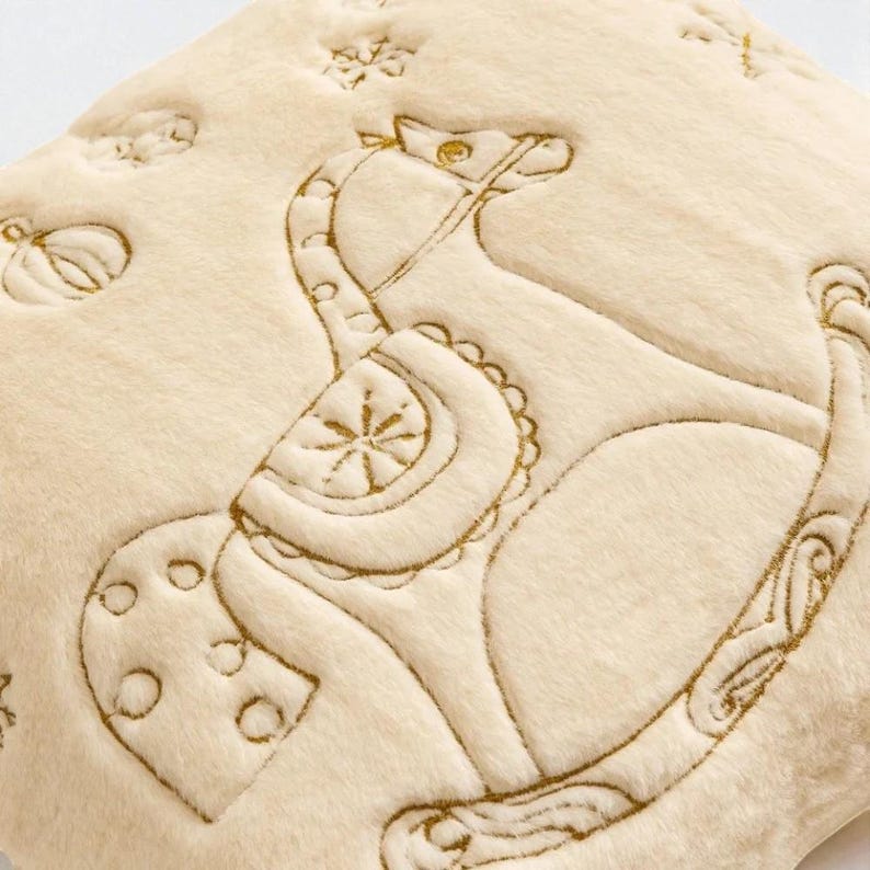 Pu&ograve; includere: Cuscino in peluche color crema con un design di cavallo a dondolo ricamato con filo dorato. Il cuscino presenta ulteriori dettagli ricamati di una zucca, stelle e altre forme. Il materiale in peluche offre un aspetto morbido e strutturato.