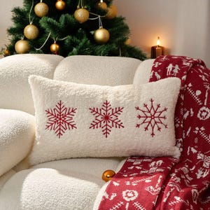 Può includere: Un cuscino rettangolare bianco con tre fiocchi di neve rossi ricamati. Il cuscino è su un divano bianco, con una coperta a fantasia rossa e bianca e un albero di Natale sullo sfondo. Sono visibili anche una candela accesa e ornamenti dorati.