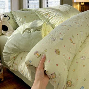 Op de afbeelding: Een lichtgroene beddengoedset met een grillig patroon van kleine cartoonfiguren, harten en andere vormen. De set bevat een dekbedovertrek, kussenslopen en een lang bolsterkussen. Een knuffelhond is zichtbaar op het bed.