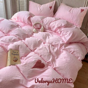 Puede incluir: Un juego de cama rosa con una colcha y fundas de almohada texturizadas. Las fundas de almohada tienen pequeños detalles de corazón. Un libro titulado "Ver a Espana" y unos auriculares blancos están en la cama. El texto "VeloryaHOME" está en la parte inferior.