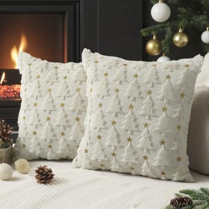 Può includere: Due cuscini decorativi color crema con un motivo a trama di alberi di Natale e stelle dorate. I cuscini sono di forma quadrata e hanno un aspetto morbido e soffice, perfetti per l'arredamento festivo.