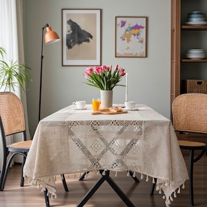 Peut inclure: Une table à manger avec une nappe beige ornée d'une dentelle décorative et de pompons. La table est dressée avec des tasses blanches, un vase de tulipes roses et une planche de service en bois. Deux chaises en osier sont de chaque côté.