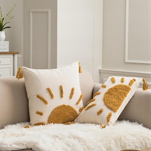 Pode incluir: Duas almofadas decorativas marfim com um desenho de sol mostarda. Cada almofada tem uma borla mostarda em cada canto. As almofadas estão em um sofá bege com uma manta branca e fofa.