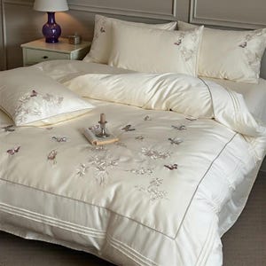 Può includere: Set di biancheria da letto color avorio con ricami di farfalle e motivi floreali. Il set include un copripiumino, federe e un cuscino decorativo. Un libro e una candela sono sul letto. La biancheria da letto ha una lucentezza sottile e un bordo plissettato.