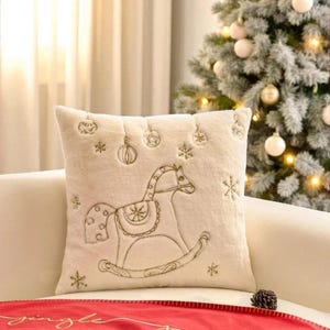 Pu&ograve; includere: Un cuscino decorativo quadrato color crema con un design di cavallo a dondolo ricamato. Il cuscino presenta fiocchi di neve, ornamenti e stelle ricamate. Il cuscino &egrave; su un divano bianco con un albero di Natale sullo sfondo.