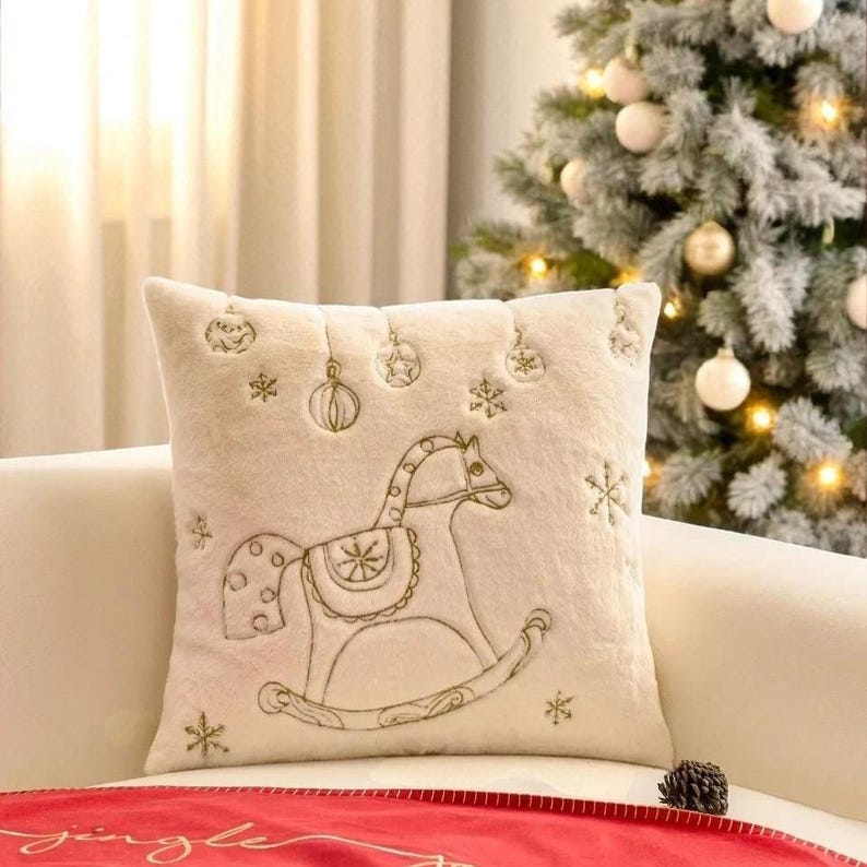 Pu&ograve; includere: Un cuscino decorativo quadrato color crema con un design di cavallo a dondolo ricamato. Il cuscino presenta fiocchi di neve, ornamenti e stelle ricamate. Il cuscino &egrave; su un divano bianco con un albero di Natale sullo sfondo.
