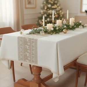 May include: A cream tablecloth with a beige reindeer pattern runner. The table is decorated with candles, greenery, and baubles, creating a festive holiday atmosphere. The table is made of wood.