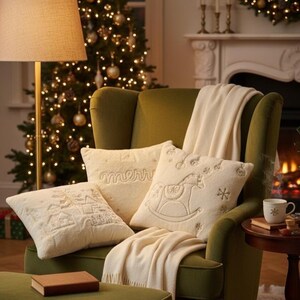 Pu&ograve; includere: Un'accogliente scena di soggiorno con una poltrona verde adornata da cuscini decorativi color crema e una coperta abbinata. I cuscini presentano disegni ricamati, tra cui un cavallo a dondolo e una scena invernale. Un albero di Natale illuminato sullo sfondo.