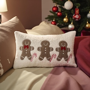 Può includere: Un cuscino rettangolare in peluche bianco con tre omini di pan di zenzero ricamati con fiocchi rossi e bastoncini di zucchero. Il cuscino è su un letto con un albero di Natale decorato sullo sfondo.