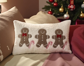Può includere: Un cuscino rettangolare in peluche bianco con tre omini di pan di zenzero ricamati con fiocchi rossi e bastoncini di zucchero. Il cuscino è su un letto con un albero di Natale decorato sullo sfondo.