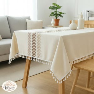 Può includere: Tovaglia rettangolare color crema con design a foglie ricamate in tonalità marroni e beige. La tovaglia ha un bordo con frange e nappe bianche. Il tavolo è in legno e ha una pianta e oggetti decorativi sopra.