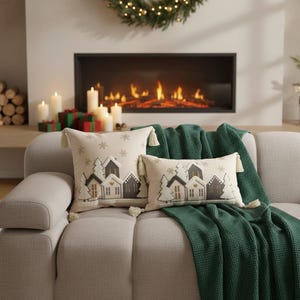 Può includere: Due cuscini decorativi con un design a villaggio invernale, con case e alberi in grigio e bianco, con dettagli a fiocco di neve. I cuscini hanno nappe color crema e sono posizionati su un divano beige con una coperta verde.