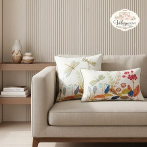Puede incluir: Dos cojines decorativos bordados con diseños de libélulas y flores sobre un sofá beige. Los cojines tienen un fondo blanco con detalles en verde, azul, naranja y rojo. El sofá está colocado contra una pared a rayas.