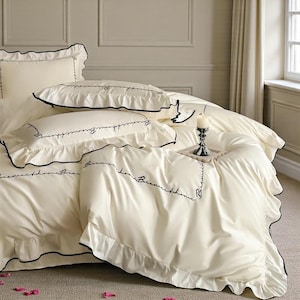 Può includere: Set di biancheria da letto color crema con finiture nere e scritte ricamate. Il set include un copripiumino e federe con bordi a balze. Una candela accesa è sul letto, aggiungendo un tocco di eleganza all'arredamento della camera da letto.