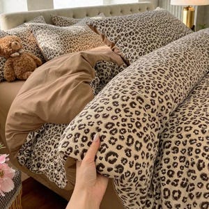 Könnte beinhalten: Ein Bett mit beigem Kopfteil und Bettwäsche. Die Bettwäsche umfasst eine braune Bettdecke, Kissen mit Leopardenmuster und ein langes Nackenkissen. Ein brauner Teddybär sitzt auf dem Bett. Der Leopardenprint hat schwarze Flecken auf beigem Hintergrund.