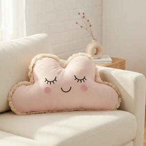 Puede incluir: Un cojín decorativo rosa con forma de nube con ojos cerrados bordados, mejillas rosadas y una boca sonriente. El cojín tiene un borde con flecos y está colocado en un sofá blanco. Es un artículo decorativo suave y afelpado.