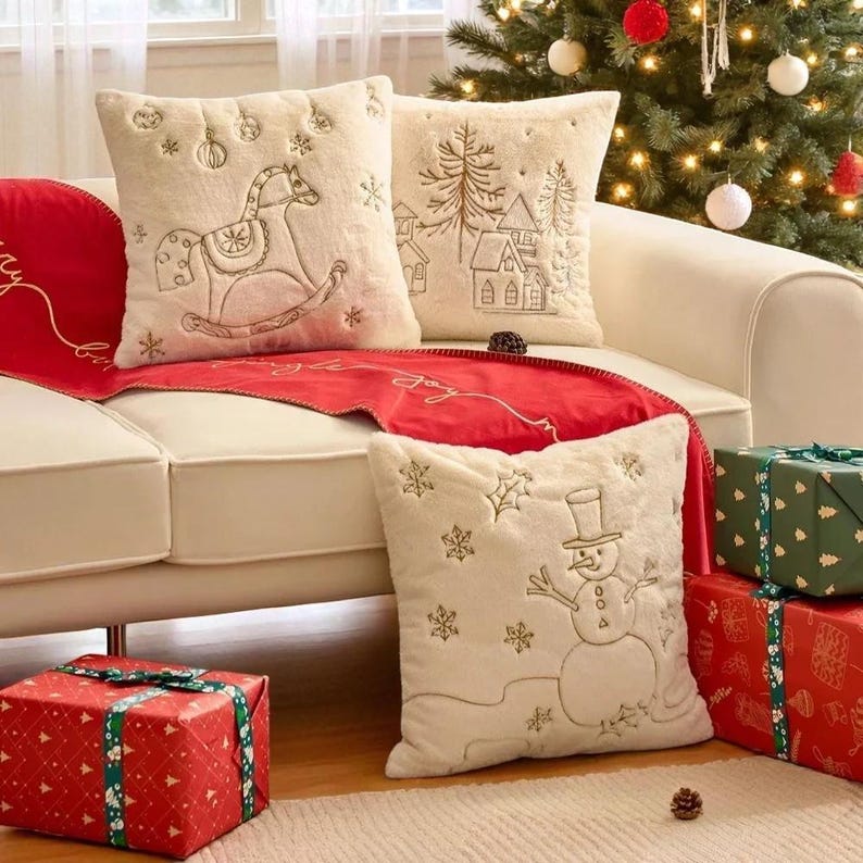 Pu&ograve; includere: Cuscini decorativi color crema con ricami a tema festivo, tra cui un cavallo a dondolo, una scena invernale e un pupazzo di neve. Un runner da tavolo rosso con la parola "joy" &egrave; drappeggiato sul divano. I regali confezionati sono posizionati nelle vicinanze.