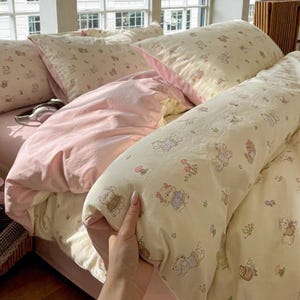 Può includere: Un letto con un piumino e cuscini rosa chiaro. La biancheria da letto presenta un motivo color crema di orsetti e fiori. È visibile anche un lungo cuscino con lo stesso motivo di orsetti.