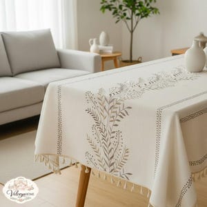 Op de afbeelding: Crèmekleurig rechthoekig tafelkleed met een botanisch ontwerp in bruin en grijs. Het tafelkleed heeft een gestippelde rand en kwastjes langs de randen. De tafel staat in een lichte kamer met een bank en decoratieve vazen.