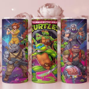 May include: A cylindrical tumbler featuring a colorful graphic print of Teenage Mutant Ninja Turtles characters, including Leonardo, Bebop, Rocksteady, Shredder, Krang, and April O'Neil, set against a brick wall and sewer background with vibrant purple and green accents.