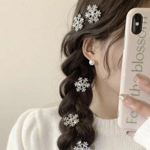 Puede incluir: Primer plano de una trenza adornada con varias pinzas para el pelo plateadas con forma de copo de nieve. Las pinzas están incrustadas con pequeños cristales brillantes. El pelo es castaño oscuro y la persona lleva un jersey blanco.