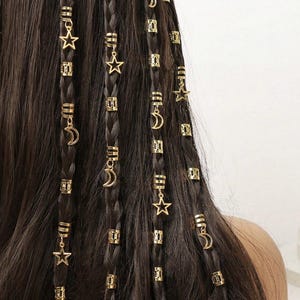 May include: Dark brown braided hair decorated with gold-coloured hair cuffs and charms. The charms feature stars and crescent moons, adding a celestial touch. The hair accessories are evenly spaced along the braids.