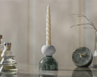 Op de afbeelding: Een decoratieve kandelaar met een witte, gedraaide kaars. De houder heeft een witte marmeren bol bovenop een groene marmeren voet. Andere decoratieve items zijn glazen flessen en een vaas met een takje.