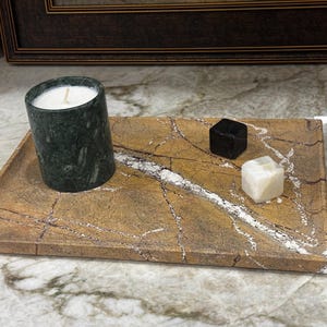 May include: A dark green marble candle holder with a lit white candle sits on a rectangular stone tray. Two small stone cubes, one black and one white, are also on the tray. The tray has a brown and white marbled pattern.