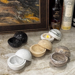 Puede incluir: Una colección de recipientes esféricos de mármol en varios colores, incluyendo negro, blanco, beige y gris. Algunos están abiertos, revelando el interior, y uno contiene una vela encendida. Los recipientes se exhiben sobre una superficie de mármol.
