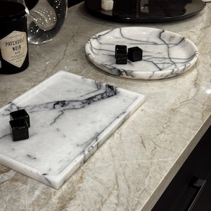 May include: A collection of marble trays and dishes with black accents. The trays are rectangular and oval, featuring white marble with gray veining. Small black square objects are placed on the trays. A black jar with white text is also visible.
