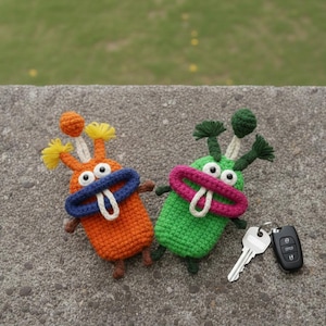 Könnte beinhalten: Zwei gehäkelte Monster-Schlüsselanhänger in Orange und Grün, jeweils mit großen Augen, einem blauen oder rosa Mund und einer weißen Zunge. Das orangefarbene Monster hat gelbe und orangefarbene Antennen, während das grüne Monster grüne Antennen hat. Ein Autoschlüssel und eine Fernbedienung liegen neben dem grünen Monster.
