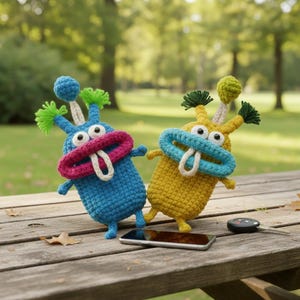 May include: Two handmade crochet monster toys in blue and yellow, with large mouths and antenna-like features. The blue monster has a pink mouth, while the yellow monster has a blue mouth. They are on a wooden table with a phone and car key.