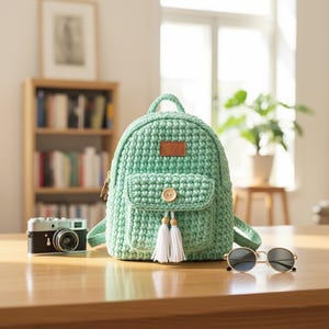 May include: A mint green crochet backpack with a front pocket and a wooden button closure. The backpack has white tassel accents and a small brown leather tag. A vintage camera and sunglasses sit nearby.