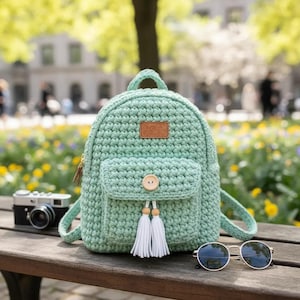 May include: A mint green crochet backpack with a small front pocket, wooden button, and white tassel accents. The bag has a small leather patch and a top handle. A vintage camera and sunglasses sit nearby on a wooden bench.