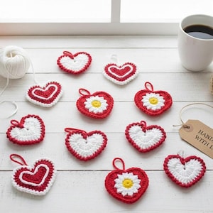 Puede incluir: Una colección de adornos de corazón de ganchillo hechos a mano en rojo y blanco, algunos con detalles de margaritas. Los corazones están dispuestos sobre una superficie de madera blanca, con una taza de café y una etiqueta que dice "HECHO A MANO CON AMOR".