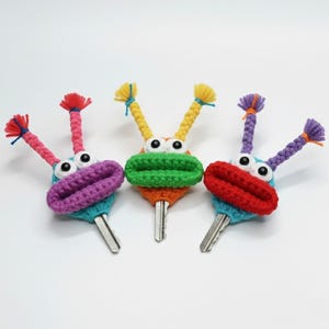 Puede incluir: Tres cubiertas de llaves hechas a mano y caprichosas. Cada cubierta presenta una cara de ganchillo con ojos saltones grandes, una boca colorida y antenas de hilo. Las cubiertas de llaves son en tonos de morado, rojo, verde, azul y naranja.