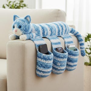 May include: A blue and white striped crochet cat-shaped organizer with three pockets. The cat has blue eyes, a pink nose, and white paws. The pockets are holding a phone, glasses, and another phone. The organizer is draped over the arm of a beige sofa.