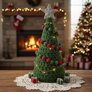 Peut inclure: Un sapin de Noël en crochet vert fait à la main, orné de pompons rouges et verts et surmonté d'une étoile argentée. L'arbre est décoré de guirlandes lumineuses et repose sur un napperon en dentelle blanche, entouré de petits coffrets cadeaux.