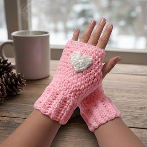 Op de afbeelding: Roze gehaakte vingerloze handschoenen met een witte hartapplicatie. De handschoenen zijn gemaakt van zacht, getextureerd garen. Ideaal om de handen warm te houden en tegelijkertijd de vingers vrij te laten. Perfect voor koud weer.