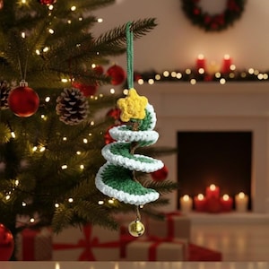 May include: A handmade crochet Christmas tree ornament. The green tree has white trim and a yellow star at the top. A small gold bell hangs from the bottom. The ornament is hanging on a Christmas tree with red ornaments and lights.