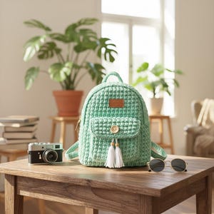 May include: A mint green crochet backpack with a small front pocket, wooden button, and white tassel accents. The backpack has a small brown leather tag. A vintage camera and sunglasses sit on a wooden table.
