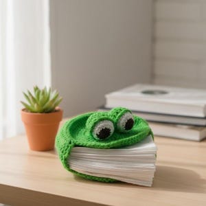 May include: A green crocheted frog tissue box cover with large, expressive eyes. The cover sits on a stack of white tissues, adding a whimsical touch to any room. A small succulent in a terracotta pot is to the left.
