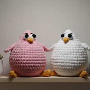 May include: Two handmade crochet bird figures. One is pink, the other is white, both with large black eyes, yellow beaks, and yellow feet. They are holding hands and sitting on a wooden shelf. The birds are a whimsical home decor item.