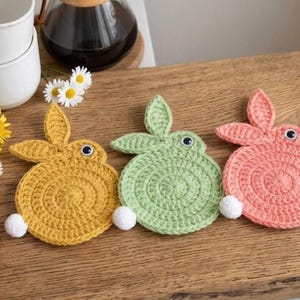 May include: Three handmade crochet bunny coasters in yellow, green, and pink. Each coaster features a round body, bunny ears, and a white pom-pom tail. The coasters are placed on a wooden surface, with daisies, strawberries, and coffee cups in the background.