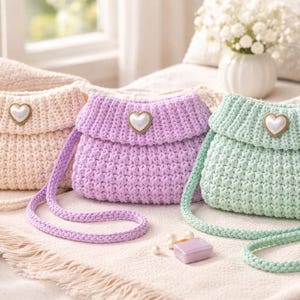 May include: Three handmade crochet purses in pastel colors: cream, lavender, and mint green. Each purse features a heart-shaped clasp and a long strap. The purses are displayed on a soft, textured surface, with a candle and flowers in the background.