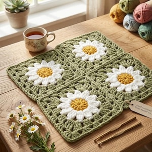 Könnte beinhalten: Vier gehäkelte Gänseblümchen-Quadrate, die zu einem größeren Quadrat angeordnet sind. Jedes Quadrat zeigt eine weiße Gänseblümchen mit gelber Mitte auf grünem Hintergrund. Häkelnadeln und Wollknäuel sind ebenfalls zu sehen.
